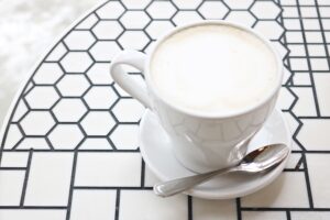 A coffee cup with cappuccino on a geometric ceramic table, accompanied by a spoon.