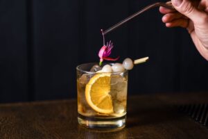 A stylish cocktail with orange slice, ice, lychee, and edible flower garnish on a dark background.