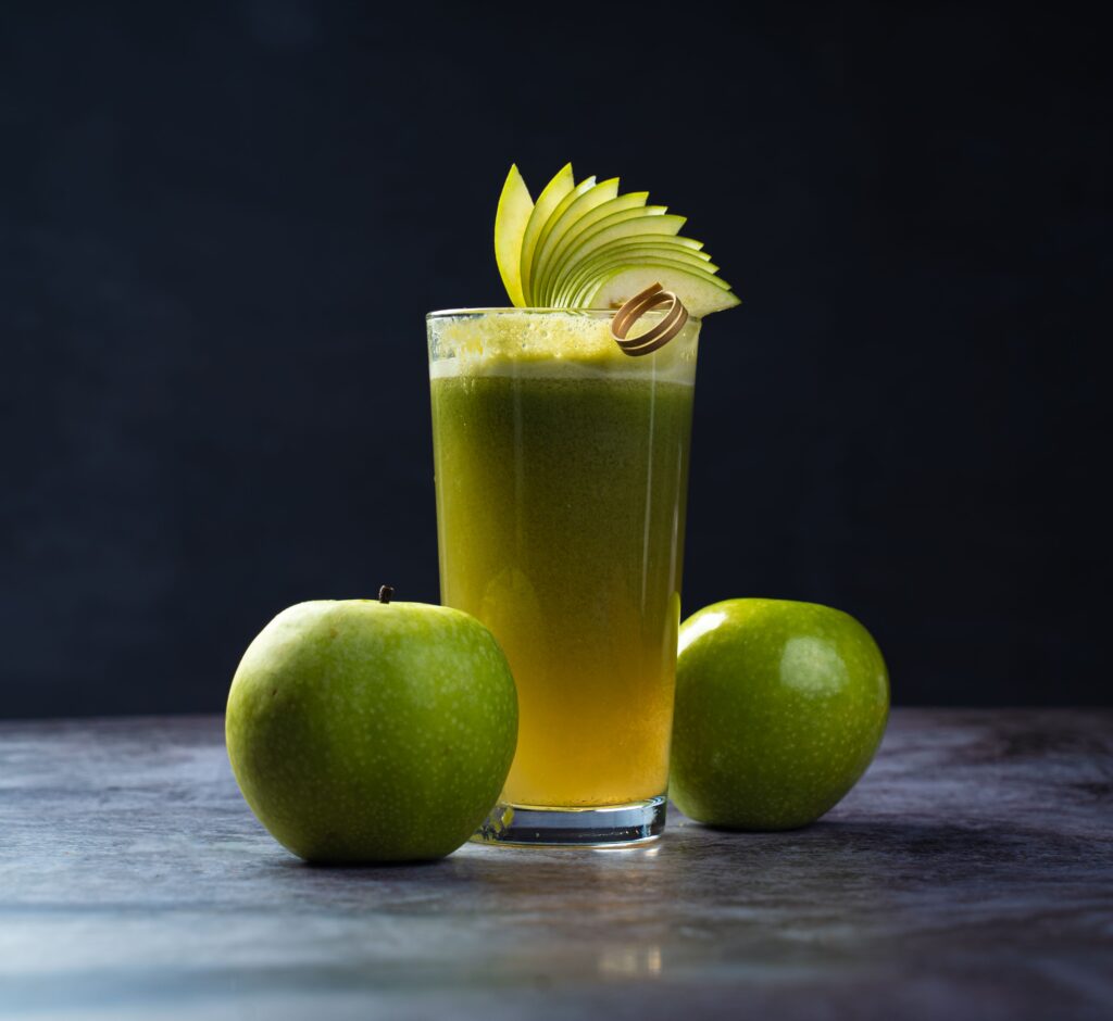 Tall glass of fresh green apple juice garnished with apple slices, accompanied by whole green apples on a dark surface.
