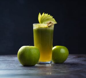 Tall glass of fresh green apple juice garnished with apple slices, accompanied by whole green apples on a dark surface.