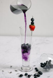 A tall glass of blueberry juice being poured, garnished with fresh berries and a strawberry.