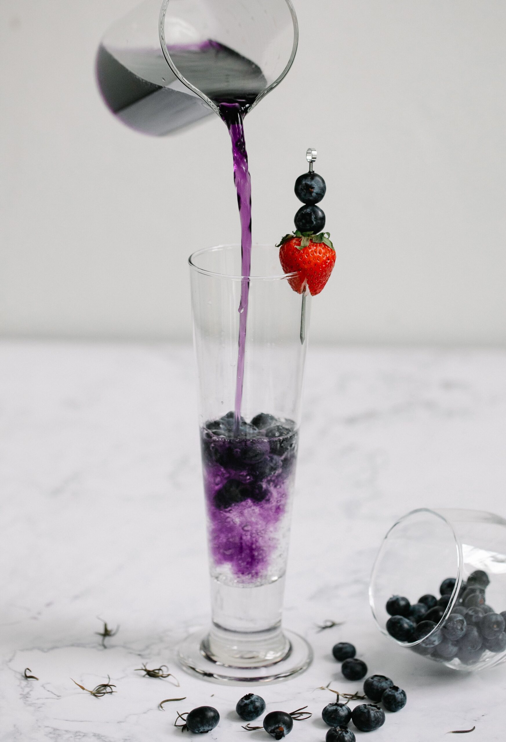 A tall glass of blueberry juice being poured, garnished with fresh berries and a strawberry.